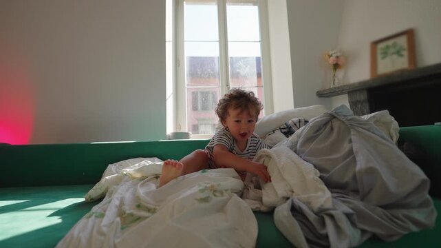 Toddler jumping onto blankets piled on sofa at home, energetic childhood action expressing carefree freedom, confidence, playful adventure and spontaneous joy