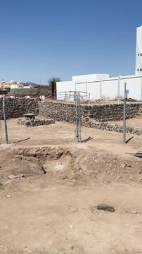 Fenced ancient stone ruins and excavation site next to modern buildings.