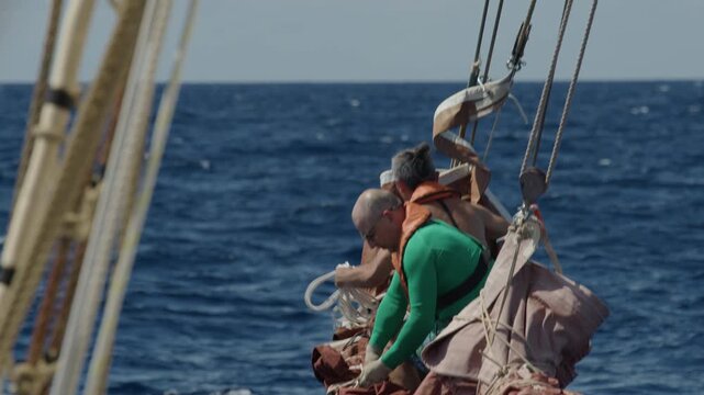 Old captain controls the crew working with the sails on the bow of the big expeditional sailing vessel moving in the rough Atlantic Ocean