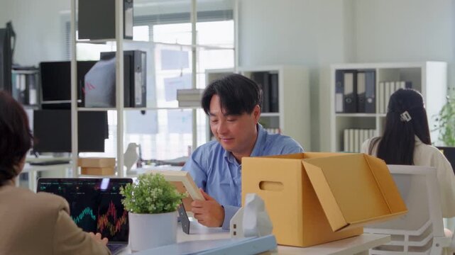 A businessman takes off his employee lanyard and ID badge while standing over a cardboard box, packing up his desk in a modern office, symbolizing job termination and career transition.