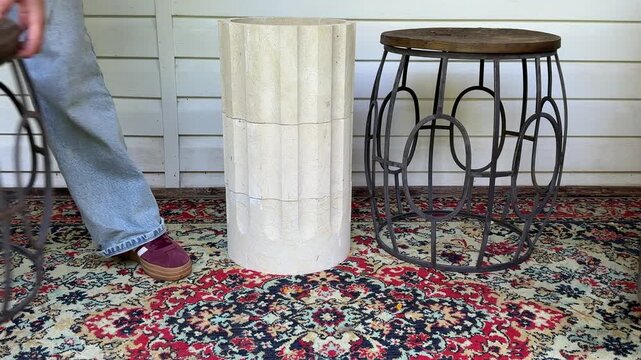 A woman compares two accent table styles a sculptural concrete pedestal and a vintage metal side table with a wooden top while styling a cozy rustic veranda. Home decorating, furniture selection
