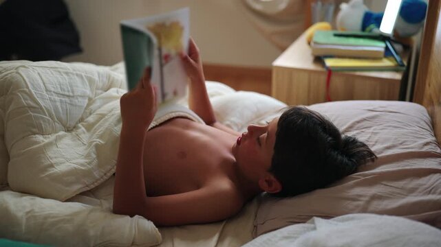 Young boy reads an illustrated story book while resting beneath a duvet in his bedroom at home, building literacy, imagination, focus and independent learning