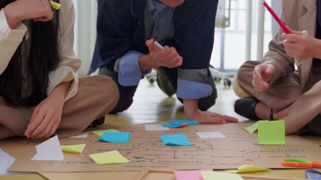 A creative team collaborates casually on the office floor, sketching a business mind map on kraft paper surrounded by colorful sticky notes, charts, and a laptop displaying data analytics.