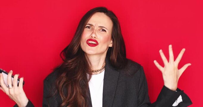 Frustrated young woman with long dark hair and red lipstick looking at smartphone screen feeling angry and stressed shouting with furious expression making hand gestures isolated on red background