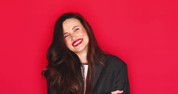 Attractive young woman with long dark hair and vibrant red lipstick laughing out loud with a happy and joyful facial expression crossing arms isolated on solid red studio background close up portrait 