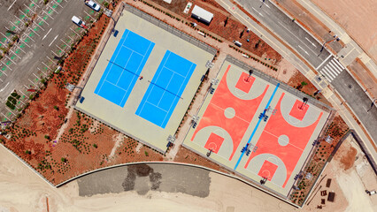 Aerial view capturing vibrant blue tennis courts and coral-colored basketball courts bordered by a parking lot and road, showcasing urban sport and recreation facilities © Nasos Zovoilis