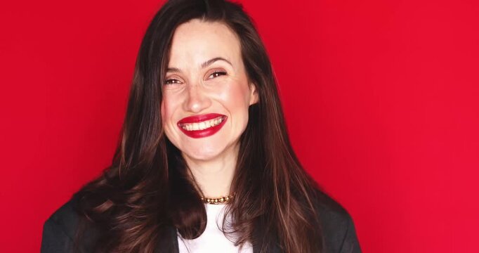 Surprised young woman with long dark hair and vibrant red lipstick spreading hands and laughing happily with a joyful and excited facial expression isolated on a solid red studio background close up