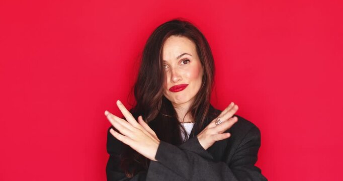 Attractive young woman with long dark hair and vibrant red lipstick showing crossed arms rejection sign with a skeptical disapproval facial expression isolated on solid red studio background close up
