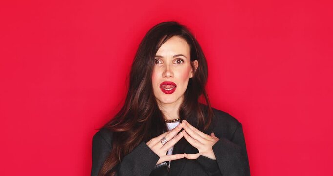 Attractive young woman with long dark hair and vibrant red lipstick rubbing hands together with a cunning sly facial expression and clever plotting smile presenting something on red studio background