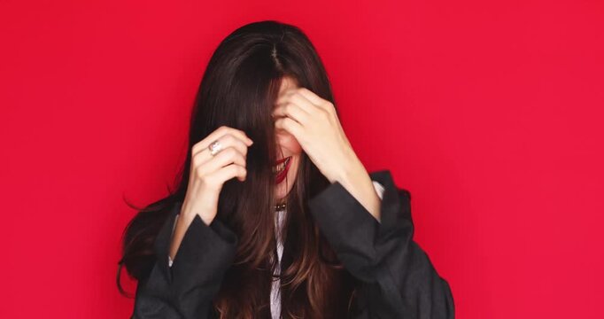 Extremely shocked surprised young woman with long dark hair and vibrant red lipstick covering mouth with hands looking at camera with wide open eyes reacting incredible news isolated on red background