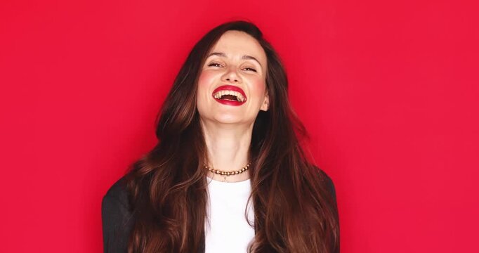 Attractive young woman with long dark hair and vibrant red lipstick smiling broadly with closed eyes and a joyful happy facial expression isolated on a solid red studio background close up portrait