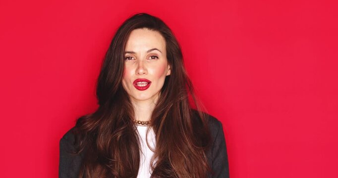 Attractive young woman with long dark hair and vibrant red lipstick looking at camera making silence gesture with finger on lips and winking playfully with a sly facial expression isolated red studio