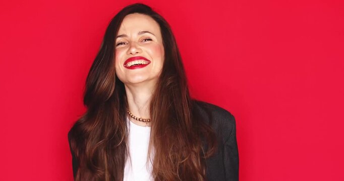 Attractive young woman with long dark hair and vibrant red lipstick making rock sign hand gestures with dynamic grimacing facial expression and happy smiling mood isolated on solid red studio