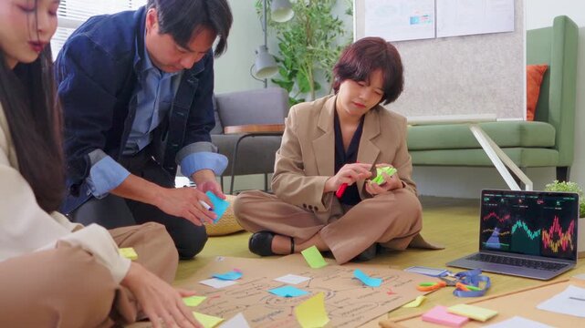 A creative team collaborates casually on the office floor, sketching a business mind map on kraft paper surrounded by colorful sticky notes, charts, and a laptop displaying data analytics.
