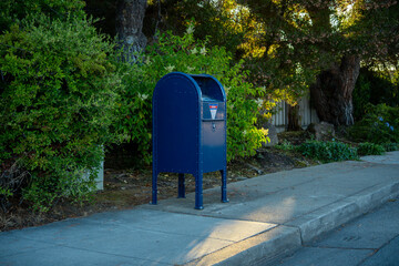 Classic blue mailbox stands on a sidewalk next to lush green bushes © Tom Harwood