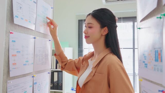 A focused professional studies technical flowcharts and software dashboards pinned to a corkboard wall, analyzing system architecture and performance metrics in a modern tech office setting.