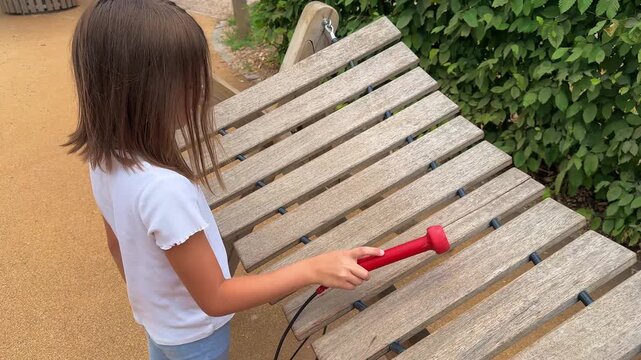 A girl in a white t-shirt plays a melody on an outdoor wooden xylophone on the playground with rubber red sticks. Musical instrument for children. High quality 4k footage