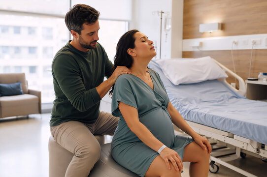 Man doing back massage for his pregnant wife sitting on the birth ball. Partner assisted birth support in hospital room. Couple during labor and childbirth preparation. 
