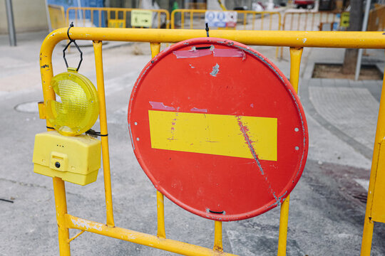 Road barrier blocks vehicle access. Red circular sign fills background. Yellow warning lamp hangs nearby. Temporary fencing surrounds repair zone. Traffic restriction protects passing workers.