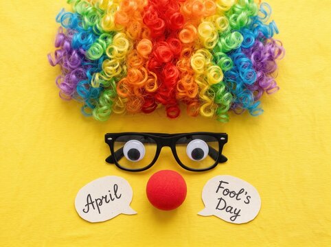 Bright rainbow colored curly clown wig with black glasses and a red foam nose on a yellow fabric background with two speech bubbles reading april fools day