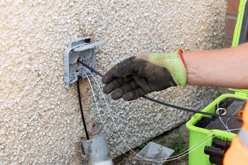 Telecoms engineer routing fibre optic drop cable through exterior wall entry bracket on pebbledash wall © Tosh Lubek