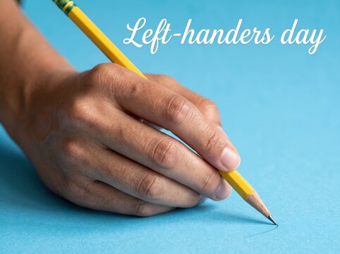 Left hand holding a yellow wooden pencil writing on a plain light blue paper surface with white text overlay reading left handers day