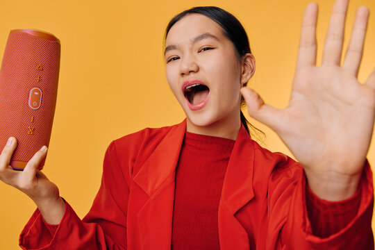 Young woman in red jacket holding wireless speaker singing or shouting on yellow background with hand raised for expression emotion and lifestyle music enjoyment.