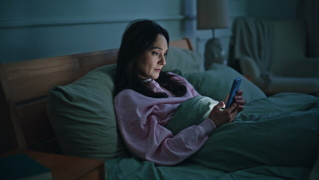Woman browsing smartphone bed spending lonely evening home. Depressed brunette