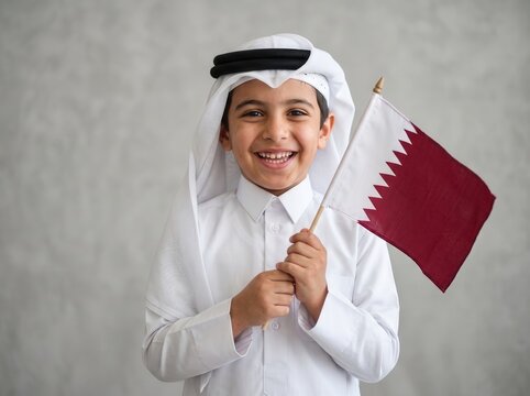 Smiling young boy wearing traditional white thobe and ghutra holding a small qatar flag against a neutral grey textured wall background