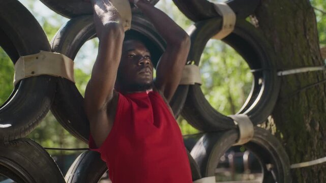 Young adult African American man performing hanging leg raises on tire wall obstacle course. Clip is ideal for fitness brand commercials, active lifestyle marketing, and gym promotions