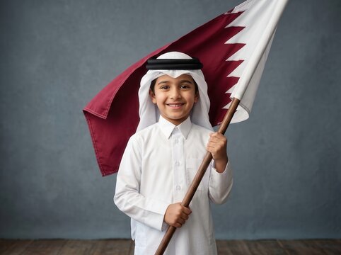 Young boy wearing traditional white thobe and ghutra smiling while holding a large flag of qatar against a textured grey background in a studio setting