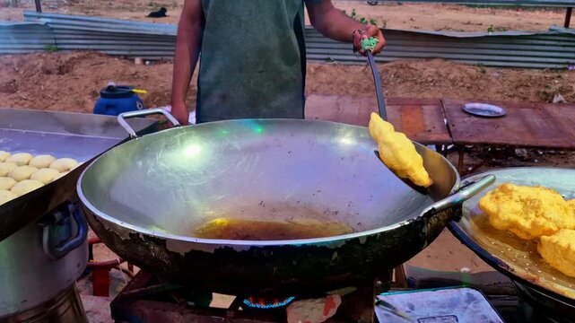 A street food vendor fries freshly rolled bhaturas in a large hot oil-filled wok over a gas burner, using a long slotted spatula. Stacks of dough balls and golden puffed puris.