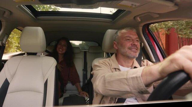 Mature man driving car while cheerful female passenger laughing in back seat