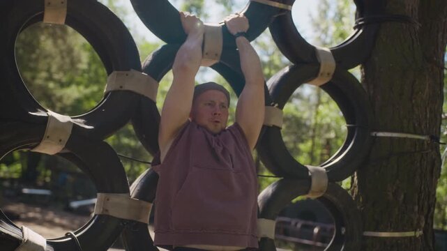 Caucasian young adult man working out abs hanging on tire obstacle course in slow motion, performing flip. Footage is ideal for promoting outdoor fitness, adventure parks, or team building