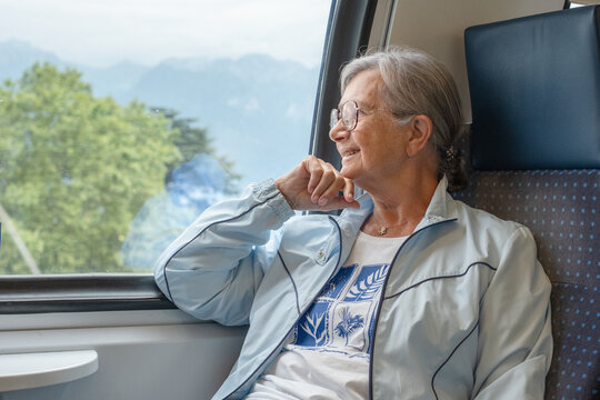 Senior woman sitting by a train window, looking out at the mountain landscape, expressing happiness and contentment while traveling