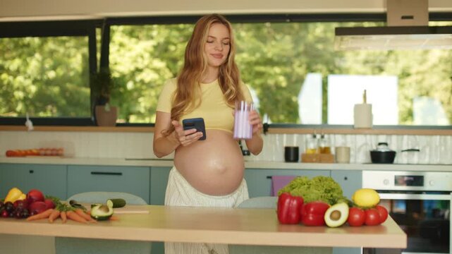 Expectant mother checking her phone while drinking glass of fresh milk in bright contemporary kitchen. Healthy pregnancy, balanced nutrition concept. 4K