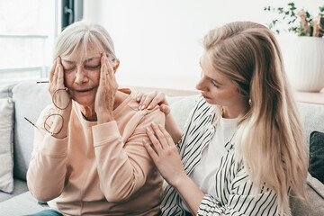 Senior woman suffering from headache or migraine, feeling painful pressure while adult daughter...