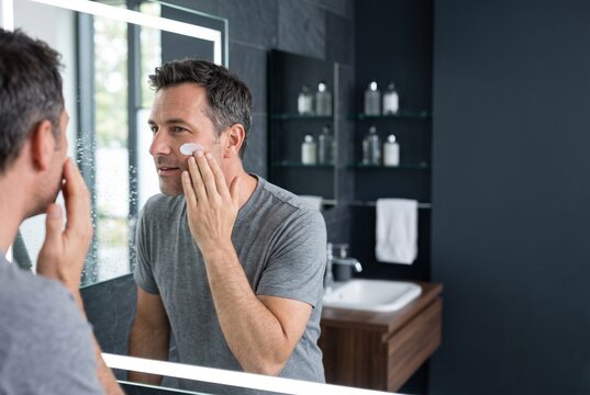 Handsome middle-aged man applying moisturizing face cream while looking at his reflection in a modern bathroom mirror, focusing on skincare and grooming routine