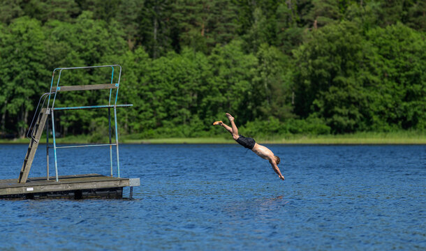 Young man finishing headfirst dive beside wooden platform on blue lake.