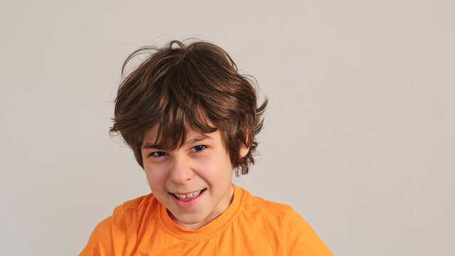 Close portrait of a smiling boy in an orange shirt making a playful face against a plain wall with copy space. Childhood emotions, confidence, and simple indoor play support family lifestyle stories.
