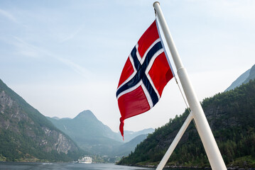 Fototapeta premium Red Norwegian flag waving on a boat with mountains and fjord water in the background on a bright summer day.
