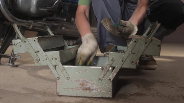 Man's gloved hands holding a wrench, selecting tools from an open metal toolbox while working on a motorcycle in a garage, focusing on maintenance and repair
