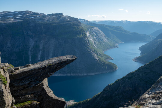 Dramatic cliff ledge above a deep Norwegian fjord valley under clear blue sky, showcasing rugged mountains and glacial lake scenery.