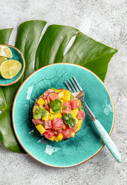 Tropical style gourmet tuna mango salad tartar with cilantro and purple onion. On blue plate, tropical leaves