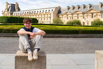 Obraz premium A smiling young man wearing eyeglasses sits on a stone block outdoors in Paris, France. A historic building and a manicured green lawn are visible in the background.
