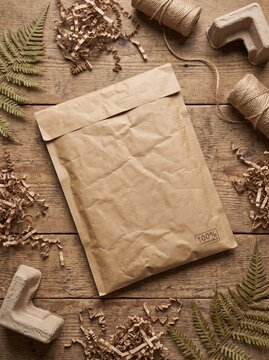Eco friendly packaging materials including a kraft paper envelope, twine, shredded paper filler, and fern leaves on a wooden background