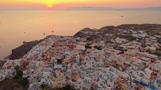 Drone view of Oia village hillside glowing in warm sunset light with blue dome churches in Santorini Greece