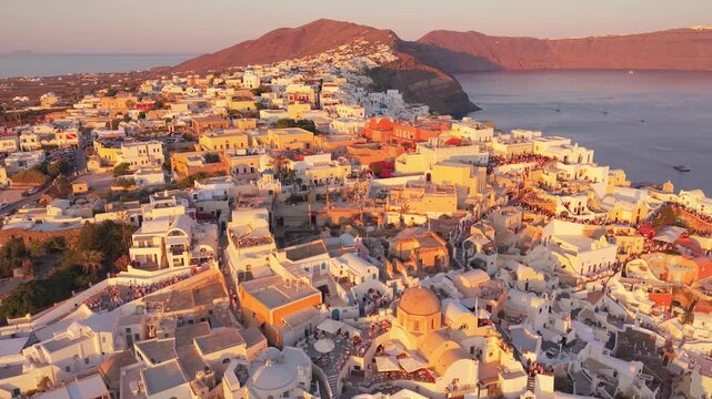Golden hour drone of Oia hillside village at sunset showcasing iconic windmills and white Cycladic architecture Santorini Greece