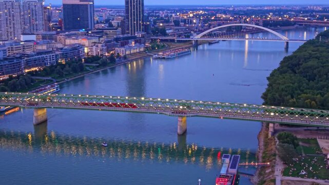 City bridge and river at night. People enjoy the riverbank while boats pass under the bridge in a city at night.