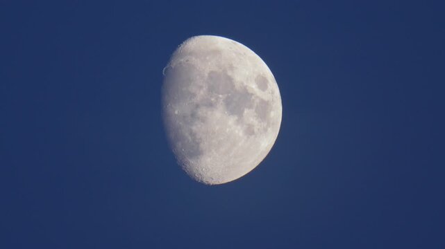 Real time 4K telephoto video of a bright waxing gibbous moon showing detailed surface craters against a deep blue twilight sky.